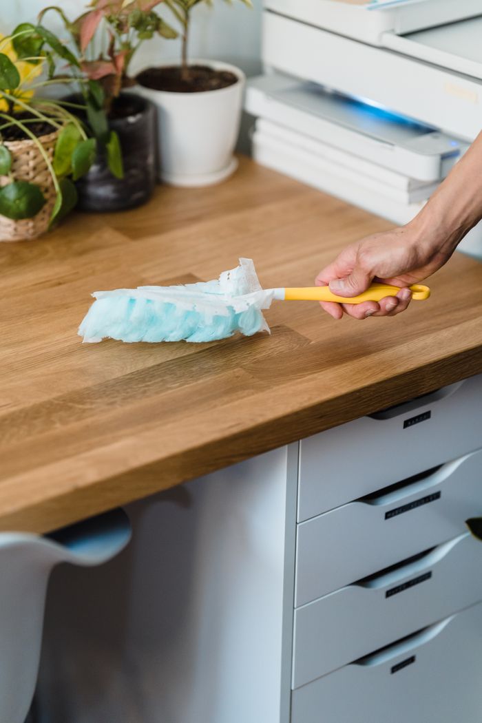 Une main utilise un plumeau pour dépoussiérer un bureau en bois, entouré de plantes et d’objets de bureau.